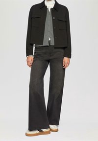 Schwarze verkürzte Jacke mit zwei Brusttaschen über einem grauen Hemd, kombiniert mit weit geschnittenen schwarzen Jeans und weißen Sneakers mit braunen Sohlen.