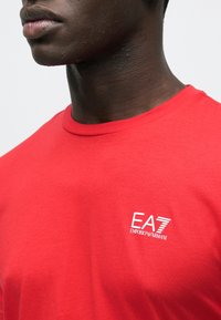 Maglietta rossa in cotone con scollo rotondo e maniche corte, con logo bianco EA7 Emporio Armani stampato sul petto. Tessuto liscio.