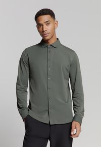 Olijfgroene blouse met lange mouwen, gemaakt van gladde stof. Beschikt over een knoopsluiting aan de voorkant, een puntige kraag en een subtiele glans.