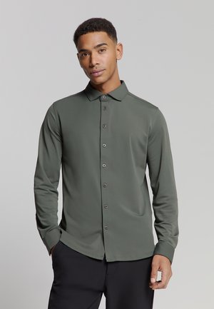 Olijfgroene blouse met lange mouwen, gemaakt van gladde stof. Beschikt over een knoopsluiting aan de voorkant, een puntige kraag en een subtiele glans.