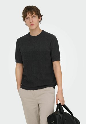 Mladý muž v čiernom krátkym rukávom svetri a béžových nohaviciach, držiaci v pravej ruke čiernu koženú tašku na rukoväti.