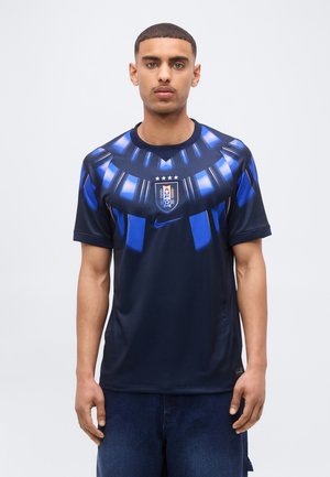 Jeune homme portant un maillot de football bleu foncé avec des motifs géométriques bleu clair et le blason de l'Association de football d'Uruguay sur la poitrine.