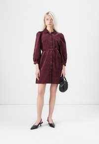 Bordeauxfarbene Hemdkleid aus strukturiertem Stoff, mit Kragen, durchgehender Knopfleiste, Taillengürtel und puffärmel. Kombiniert mit einer schwarzen Handtasche.