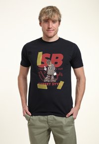 Camiseta de algodón negra con un gráfico colorido que presenta un personaje de dibujos animados en una patineta, texto en negrita y detalles de cinta amarilla.