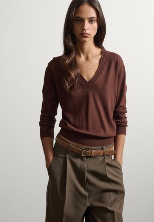 Giovane donna con lunghi capelli castani che indossa un maglione marrone a scollo a V e pantaloni oliva con vita alta e cintura beige, mani nelle tasche.