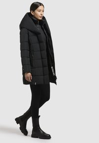 Giacca a vento nera con grande cappuccio, design trapuntato, tasche laterali con zip e etichetta "knluo", abbinata a stivaletti neri e leggings.