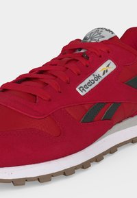 Κόκκινο αθλητικό παπούτσι Reebok με σκούρο γκρι λογότυπο στο πλάι, λευκή ενδιάμεση σόλα, καουτσουκένια εξωτερική σόλα και κόκκινες κορδόνια σε ανοιχτό φόντο.