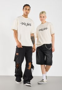 Krémové oversized trička s černým grafickým potiskem, nošená s roztrhanými černými džíny a cargo šortkami. Doplňky zahrnují řetěz a kostkované slip-on boty.