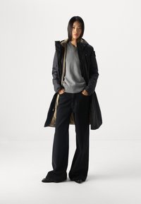 Schwarze lange Daunenjacke, grauer Strickpullover, schwarze weite Hose und schwarze Schuhe. Glatter Stoff mit einem warmen Innenfutter.