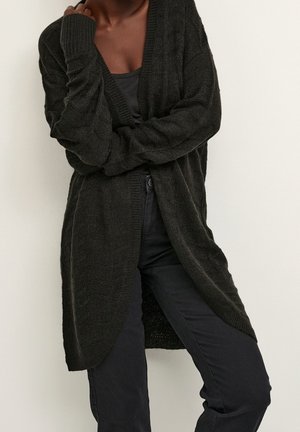 Personne portant un long cardigan noir en tricot par-dessus un top noir et un pantalon noir, debout avec un bras croisé et l'autre main près du visage.