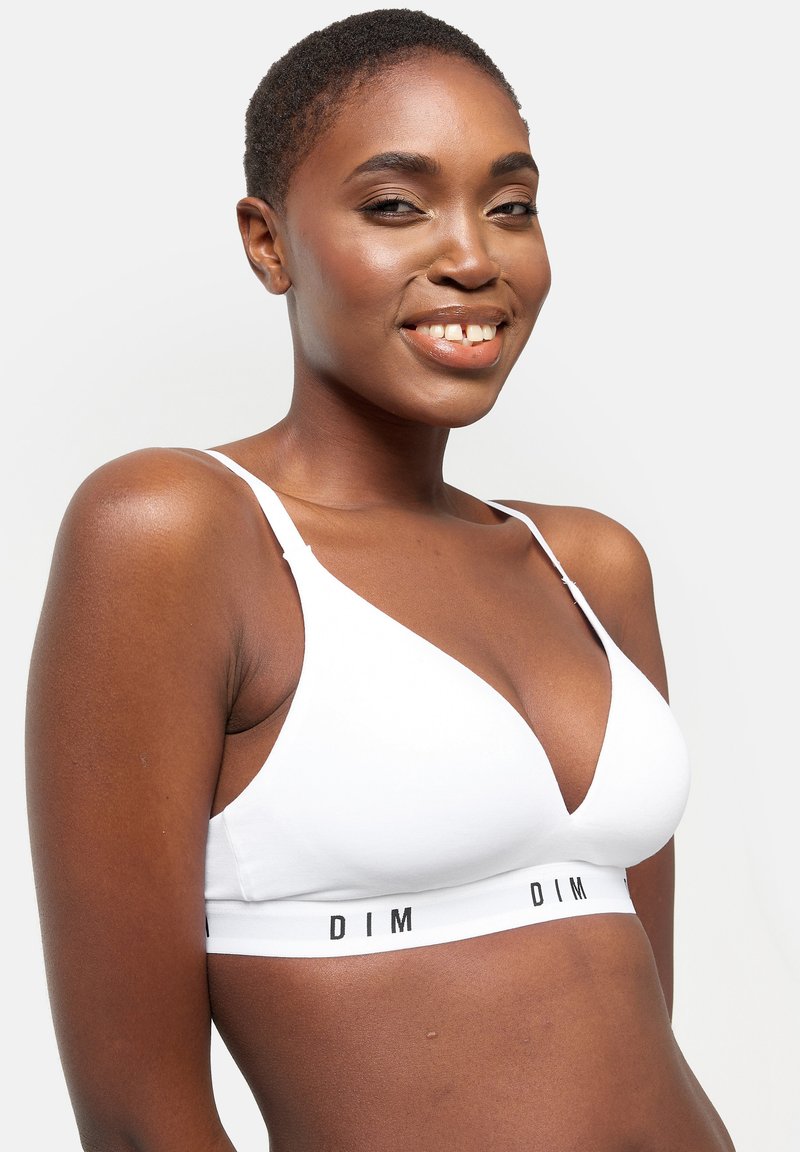 Donna sorridente con capelli corti che indossa un reggiseno bianco senza fili con una fascia elastica con il marchio "DIM" su uno sfondo bianco semplice.