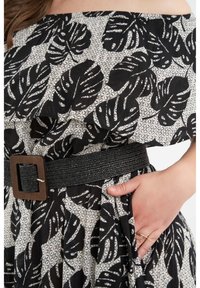 Musta ja valkoinen mekko, jossa on olkapäitä paljastava muotoilu, ja jossa on lehväkuvio sekä geometriset taustat. Mukana on leveä, teksturoitu musta vyö.