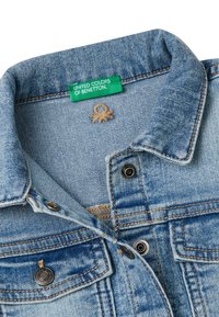 Veste en denim bleu clair avec un col large, une étiquette verte portant la mention "United Colors of Benetton" et des accents en boutons-pression argentés.
