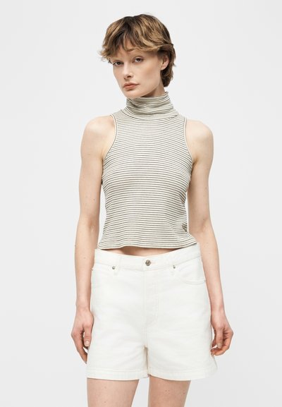 Νεαρός/ή που φοράει αμάνικο ριγέ τοπ με ψηλό λαιμό και λευκά σορτς με ψηλή μέση, στέκεται μπροστά σε απλό φόντο.