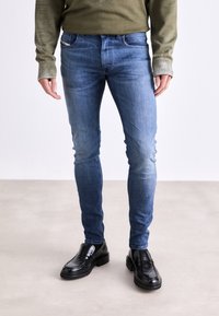Vaqueros de mezclilla azul claro con corte ajustado, combinados con un suéter verde y zapatos de cuero negros. Presentan un desgaste sutil y un diseño de cinco bolsillos.