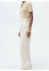 Beżowy, dzianinowy top z krótkim rękawem i falbaniastym kołnierzykiem, sparowany z białymi spodniami o wysokim stanie i szerokich nogawkach. Złote bransoletki podkreślają nadgarstek.