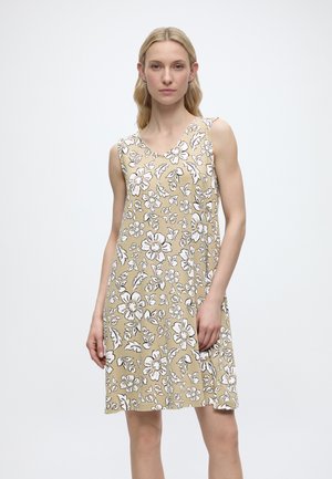 Blonde Frau steht in einem ärmellosen beigen Kleid mit weißem Blumenmuster vor hellem einfarbigem Hintergrund.