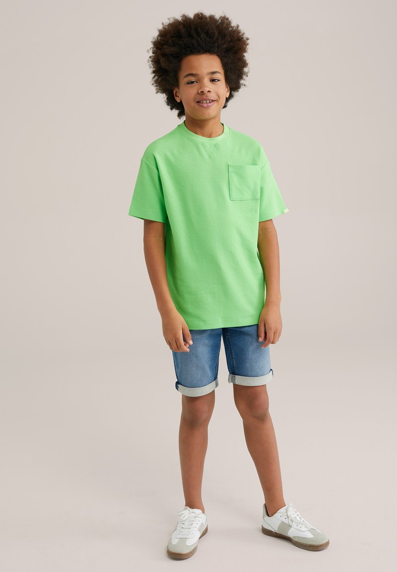 Zaļš kokvilnas T-krekls ar īsām piedurknēm un krūšu kabatu, apvienots ar sarullētām jeans šortiem un baltām sporta kedām ar pelēkiem akcentiem.