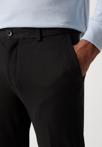 Pantalon noir en tissu texturé au design traditionnel. Doté de poches avant et d'une fermeture à bouton, présenté avec un haut à manches longues bleu.