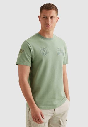 Giovane uomo che indossa una maglietta a maniche corte verde chiaro con loghi stampati e pantaloni cargo beige, in piedi con le mani nelle tasche.