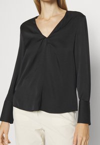 Blusa nera a maniche lunghe con scollatura a V e dettaglio annodato sul davanti; realizzata in tessuto morbido con vestibilità comoda e maniche a campana.