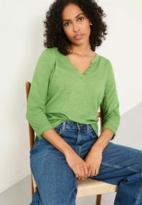 Green long-sleeve shirt with a V-neck and three buttons, paired with blue denim jeans. Shirt has a smooth texture and relaxed fit.