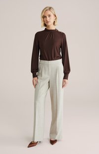 Rjava dolgoročna majica z visokim ovratnikom in gumbi, kombinirana s svetlo sivimi širokimi hlačami ter rjavimi čevlji z ostrim sprednjim delom.