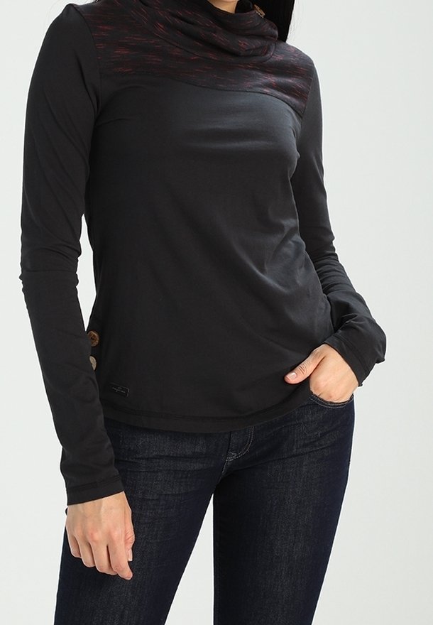 Woman wears black long-sleeve top with textured high collar and buttons on side, paired with dark blue jeans, one hand in pocket.