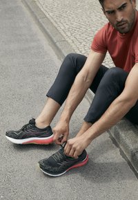 Schwarze Laufschuhe mit einer strukturierten Oberseite, pinker Zwischensohle und grünen Akzenten. Einzelne Schnürsenkel, auf einer Betonoberfläche platziert.