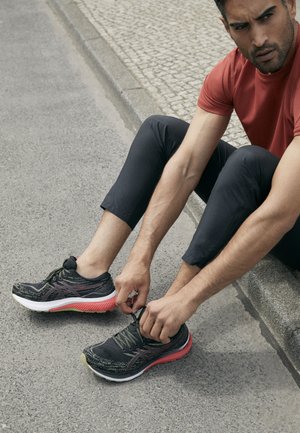 Zapatillas de correr negras con un upper texturizado, entresuela rosa y acentos verdes. Cuerdas para atar de forma individual, apoyadas sobre una superficie de hormigón.