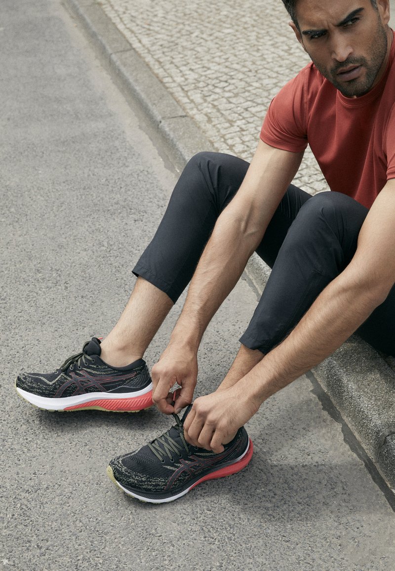 Schwarze Laufschuhe mit einer strukturierten Oberseite, pinker Zwischensohle und grünen Akzenten. Einzelne Schnürsenkel, auf einer Betonoberfläche platziert.