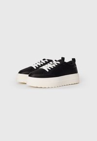 Sneaker in pelle nera con una spessa suola in gomma bianca, dotata di lacci bianchi, punta arrotondata e un motivo testurizzato sulla suola.