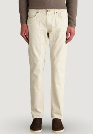 Uomo che indossa pantaloni beige a gamba dritta, maglione marrone e scarpe slip-on marrone scuro, in piedi con le mani rilassate ai lati.