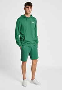 Grön hoodie med en framficka och snörning; matchas med gröna shorts. Har vita logotypdetaljer och vita sneakers.