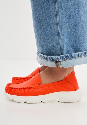 Scarpe in pelle arancione con punta arrotondata e dettagli arricciati sulla tomaia. Presentano una suola in gomma bianca spessa e pantaloni in denim blu con risvolto.