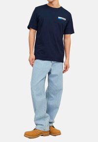 Marineblå bomulds T-shirt med et hvidt og blåt grafisk logo på venstre bryst, kombineret med lyseblå løst siddende jeans og brune støvler.
