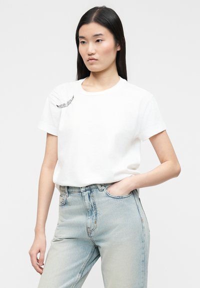 Νεαρή γυναίκα που φοράει λευκό t-shirt με σχέδιο φτερού πουλιού στον αριστερό ώμο και ανοιχτό μπλε τζιν ψηλόμεσο, με το χέρι στην τσέπη.