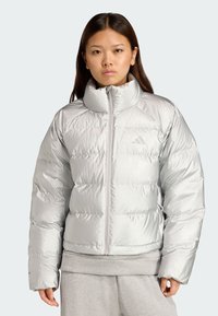 Bunda s kapucí v světle šedé barvě, vyrobená z lesklého prošívaného materiálu. Má vysoký límec, přední zip a elastické manžety.