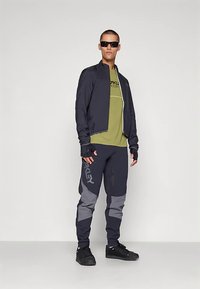 Idrottsutstyrsel med en lätt svart jacka, grön skjorta och mörkgrå byxor med logotyp. Tillbehör inkluderar solglasögon och handskar.