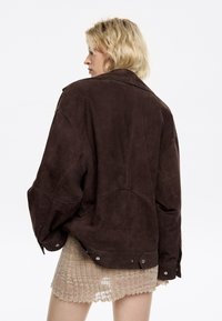 Bruine suède oversized jas met een brede kraag, naden aan de voor- en achterkant, en drukknoopaccenten aan de onderrand. Zachte textuur, losse pasvorm.