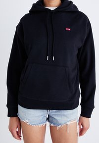 Felpa nera con cappuccio in tessuto morbido, con tasca frontale, cappuccio regolabile con cordini e punte argentate. Logo rosso sul petto. Indossata con pantaloni shorts in denim.