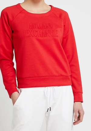 Personne portant un sweat-shirt rouge Armani Exchange avec logo en relief et pantalon blanc à cordon, debout avec une main dans la poche.