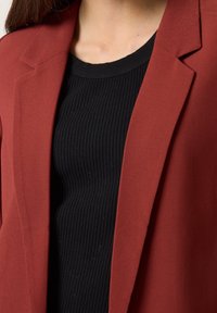 Marsalový blazer s hladkou texturou, s klopami, v kombinaci s přiléhavým černým ribbovaným topem. Zaměřte se na vzájemnou hru barev a tvarů.