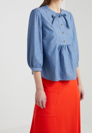 Femme portant un chemisier à manches longues bleu clair avec un nœud et des boutons, associé à une jupe orange vif avec une grande poche avant.