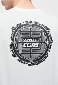 Άτομο που φοράει λευκό πουκάμισο με μαύρο κυκλικό γραφικό φρεατίου και κείμενο "CONVERSE CONS" στο πάνω μέρος της πλάτης.