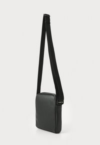 Sac à bandoulière noir avec un extérieur en cuir lisse, de forme carrée, une sangle en tissu ajustable et une fermeture éclair. Design minimaliste sans matériel visible.