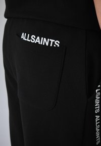 Schwarze Jogginghosen mit einer Tasche auf der Rückseite und weißem "ALLSAINTS"-Logodetail. Weiche Textur und geripptes Gewebe mit figurbetontem Schnitt.