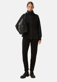 Zwarte zip-up jas met een hoge kraag, gecombineerd met zwarte strakke broek en dikke zwarte laarzen, terwijl er een gestructureerde zwarte puffy tas wordt vastgehouden.
