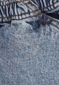 Denimstof i en falmet blå farve, med en tekstureret overflade og synlig syning i en kontrasterende orange nuance. Rynket udseende ved taljen.