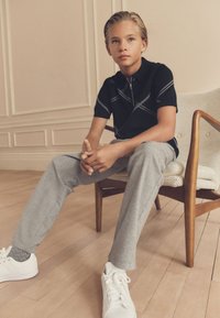 Garçon aux cheveux tirés en arrière assis sur une chaise en bois avec un coussin beige, portant une chemise noire zippée, un pantalon gris et des baskets blanches sur un sol en bois.